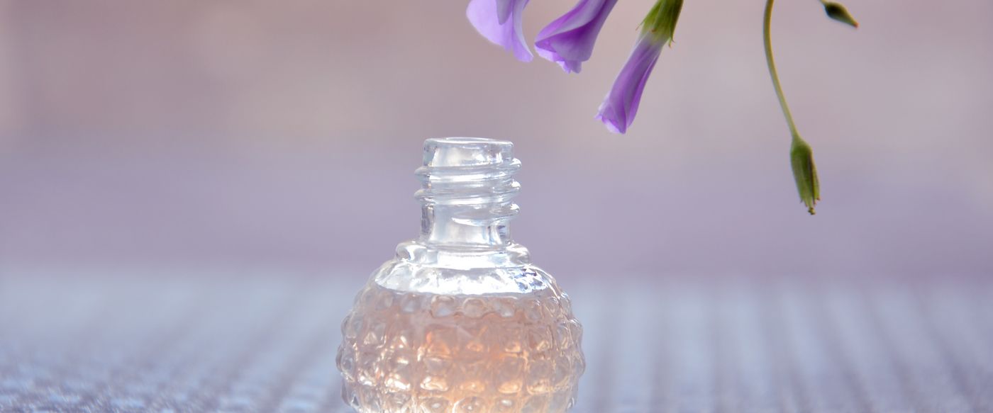 A glass perfume bottle beside purple flowers shows how fragrance in candles is built