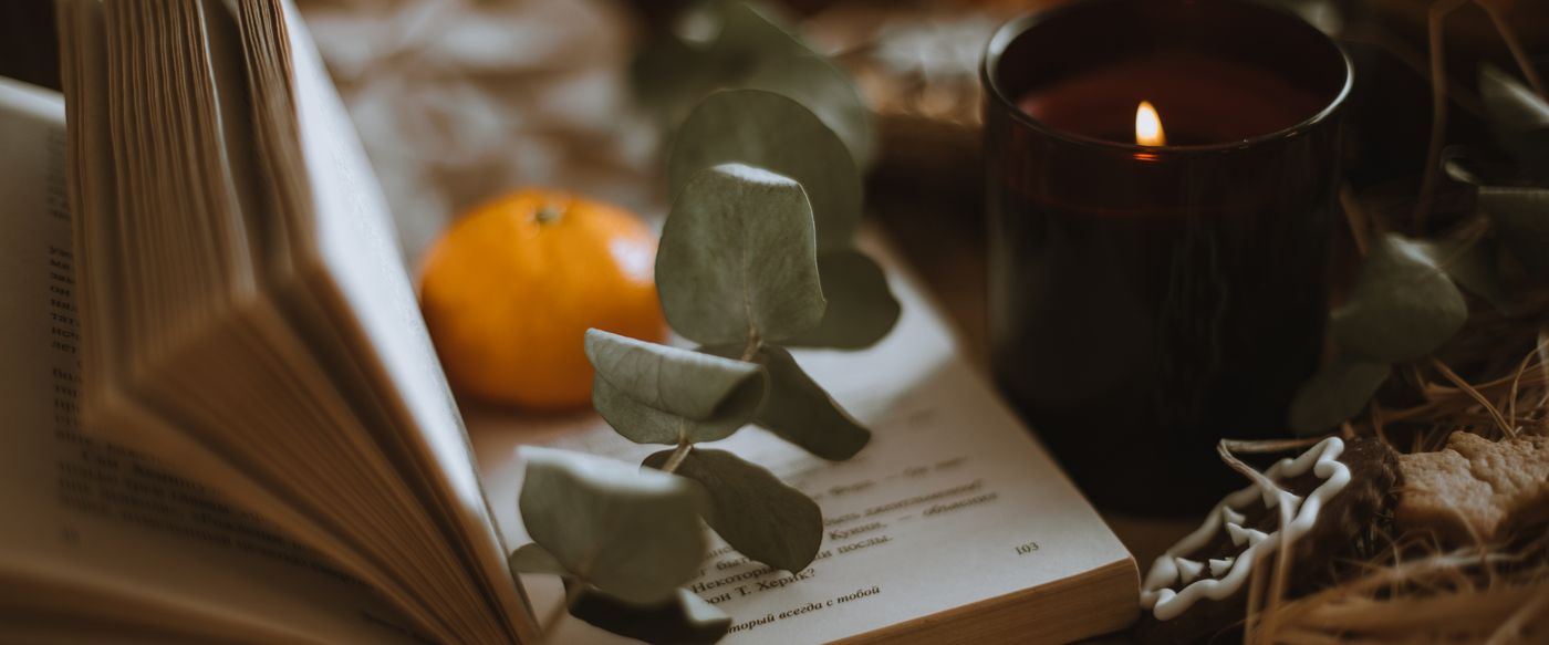 Open book, lit candle, orange and eucalyptus leaves beside vanilla scented candles