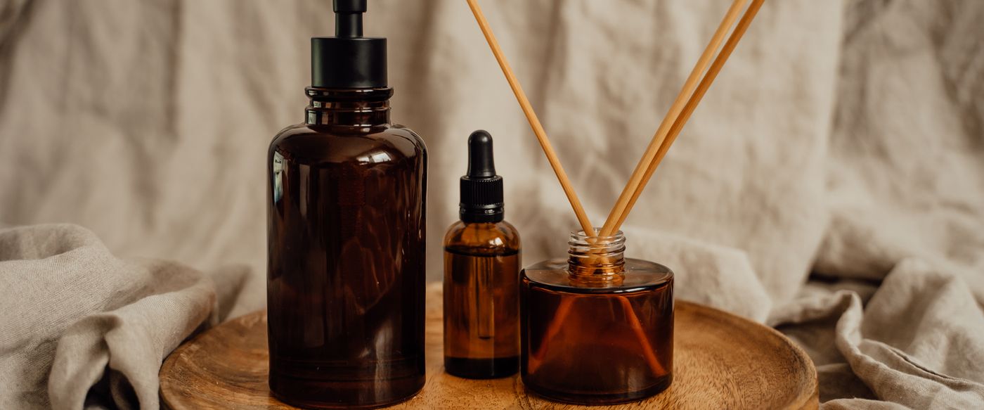 Amber glass bottles and diffuser sticks on a wooden tray behind expensive smelling candles
